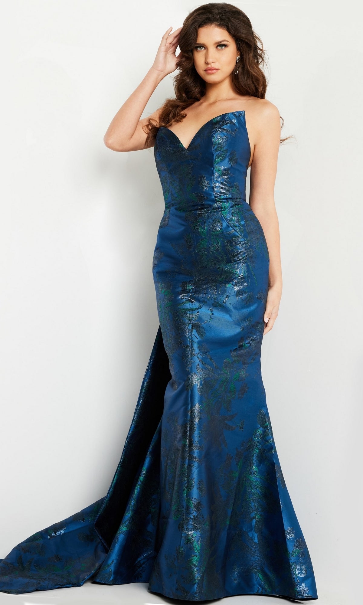 A woman stands in the Jovani 23898 Navy Blue Long Formal Dress—a strapless, floor-length mermaid gown with a slight train and metallic floral pattern—set against a plain white background.